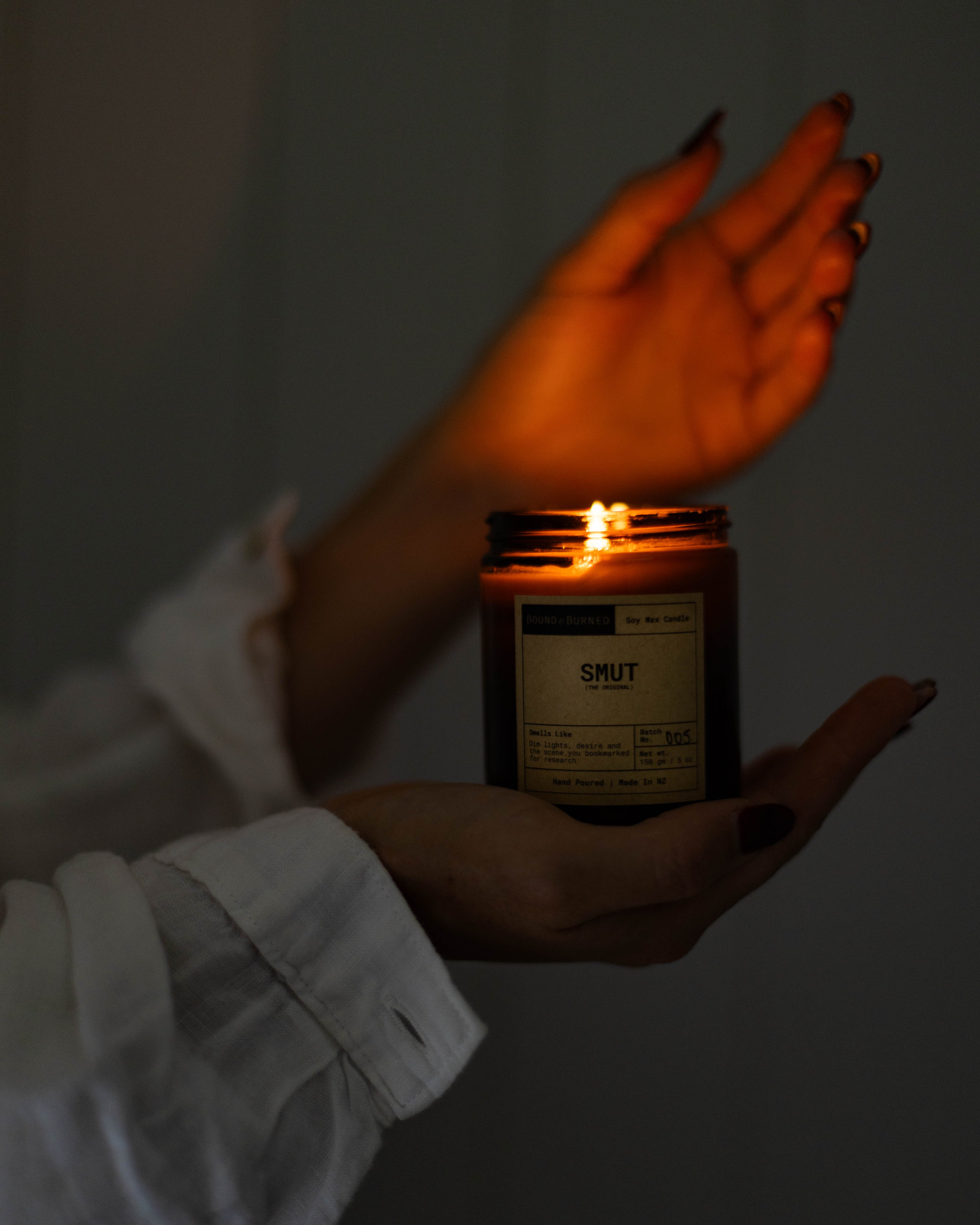 Person holding a lit candle in a jar with a label, against a dark background