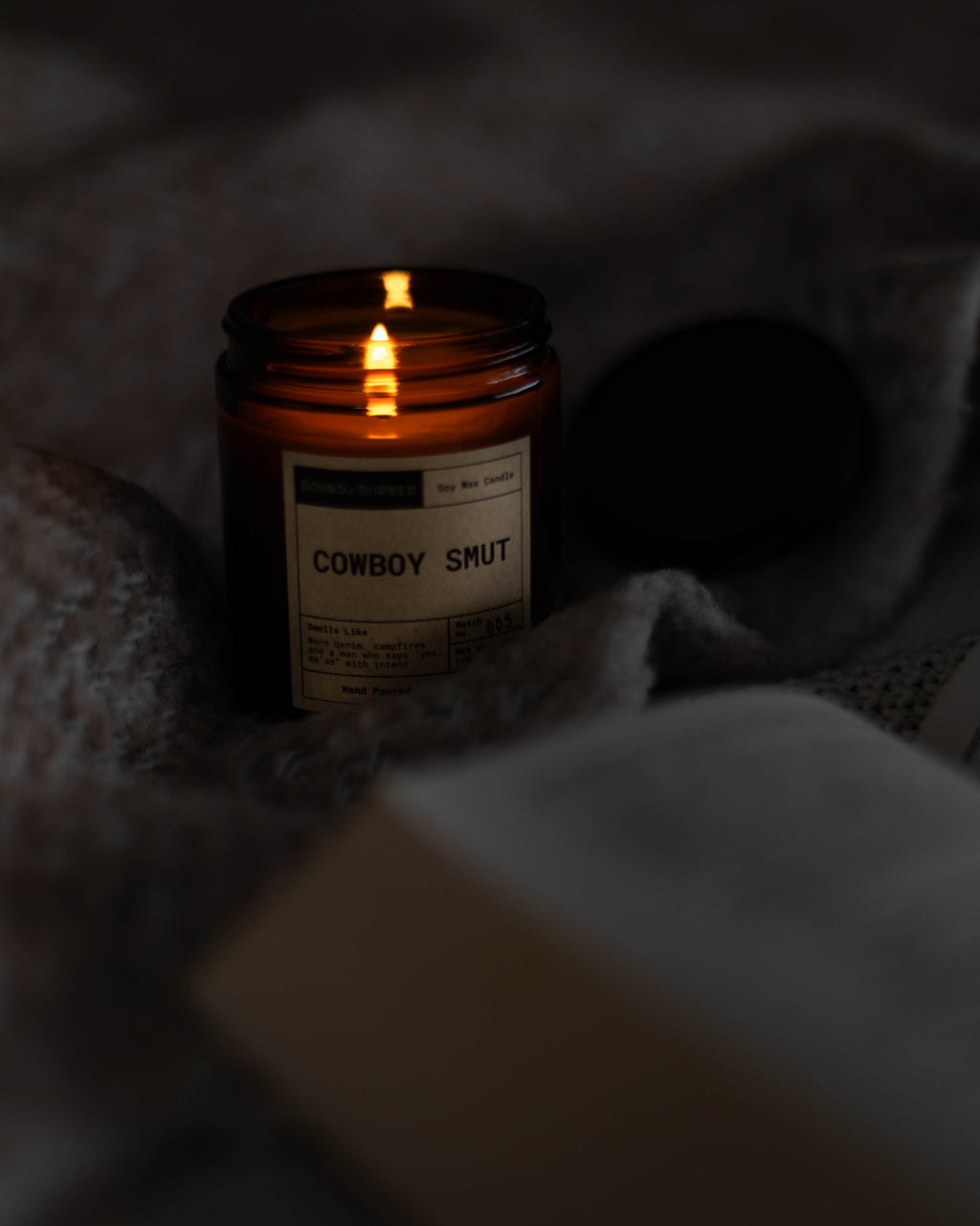 Candle labeled 'Cowboy Smut' on a textured surface with warm glow