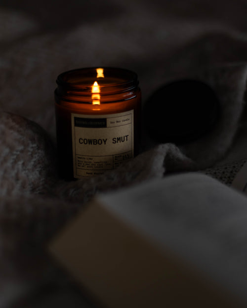 Candle labeled 'Cowboy Smut' on a textured surface with warm glow