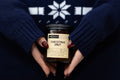 Candle labeled 'Christmas Smut' held by hands wearing dark blue knit sleeves.