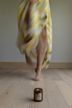 Person in a yellow and white patterned dress walking on a wooden floor with a candle in the foreground.