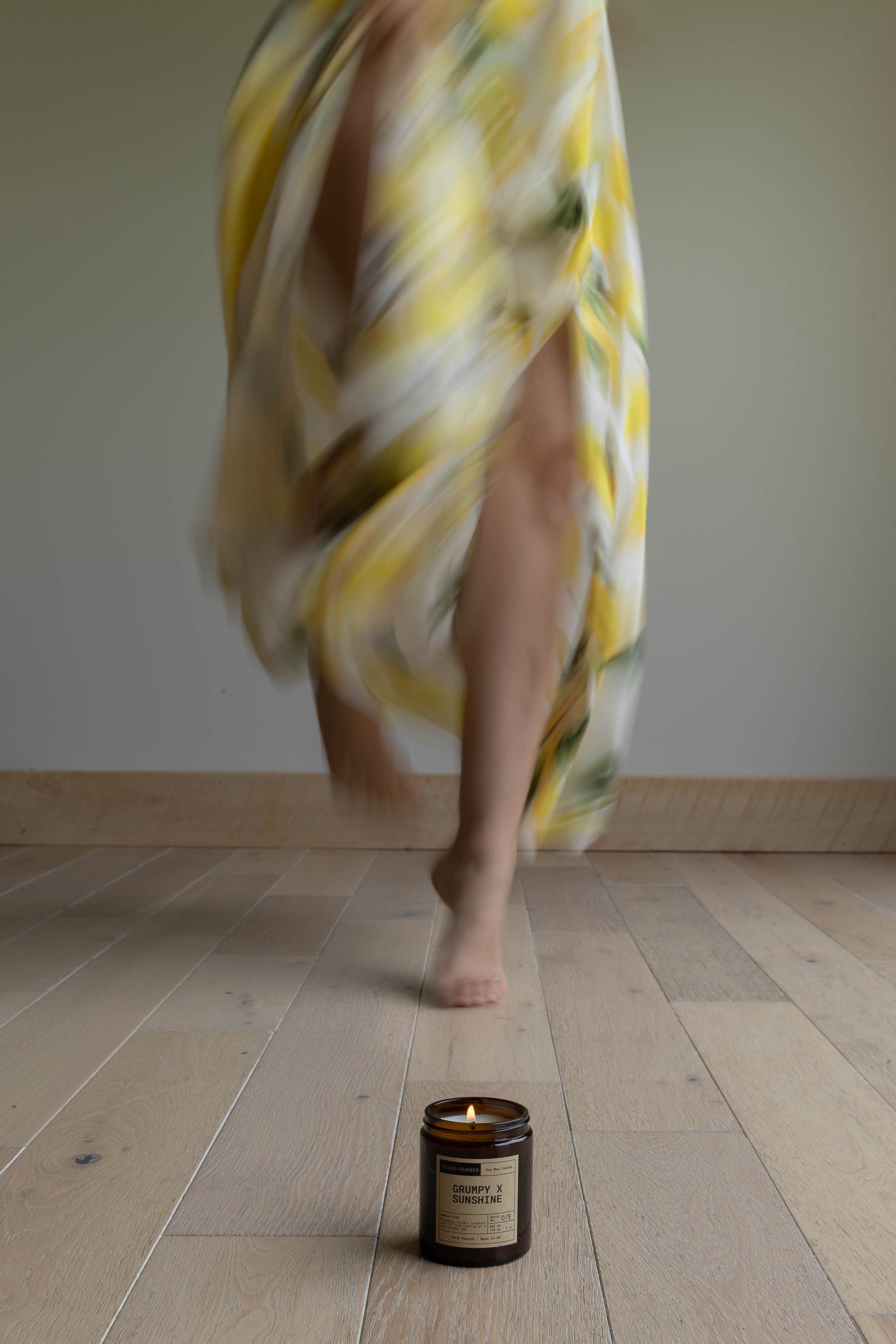 Person in a yellow and white patterned dress walking on a wooden floor with a candle in the foreground.