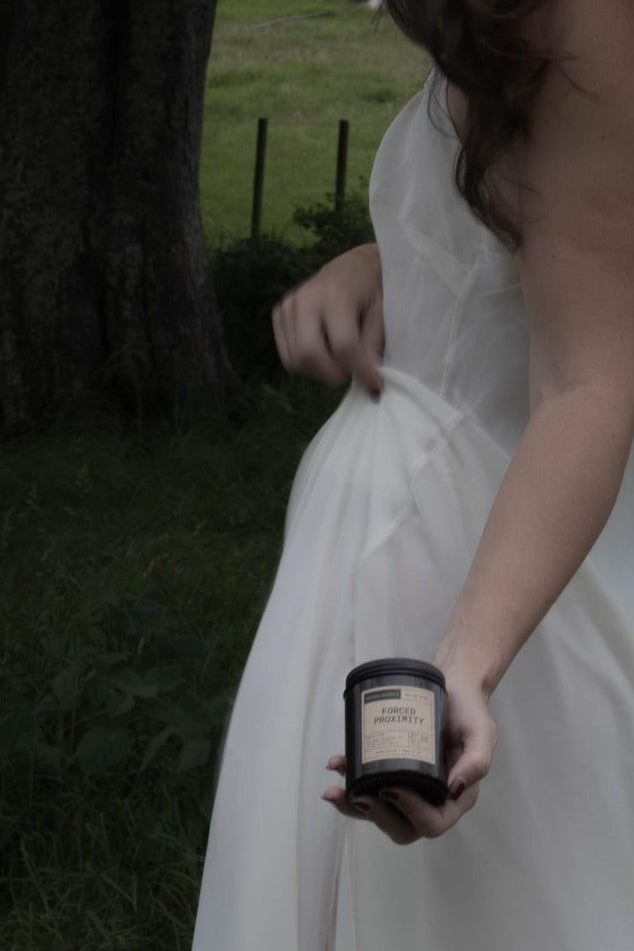 Person in a white dress holding a small candle under a tree in a grassy area