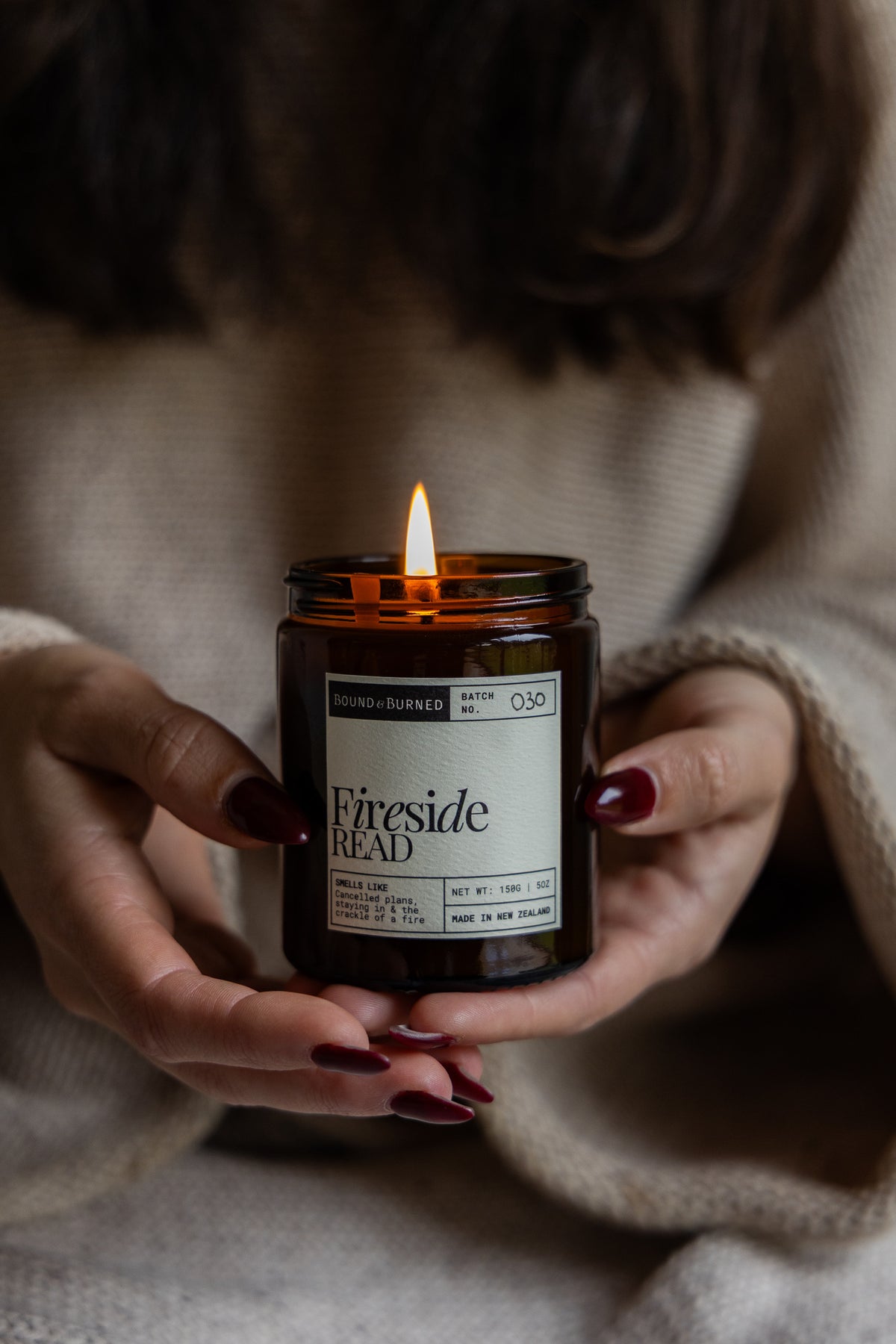 A person in a beige knit sweater holds a lit amber glass candle jar labeled 'Fireside Read' with a white label. The person's hands with dark red nail polish are visible against a cozy, softly blurred background.