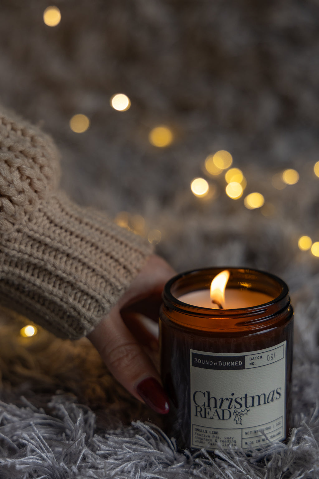 A hand in a beige knit sweater holds a lit amber glass candle jar labeled ' Christmas Read' on a gray shag rug with warm bokeh string lights in the background.