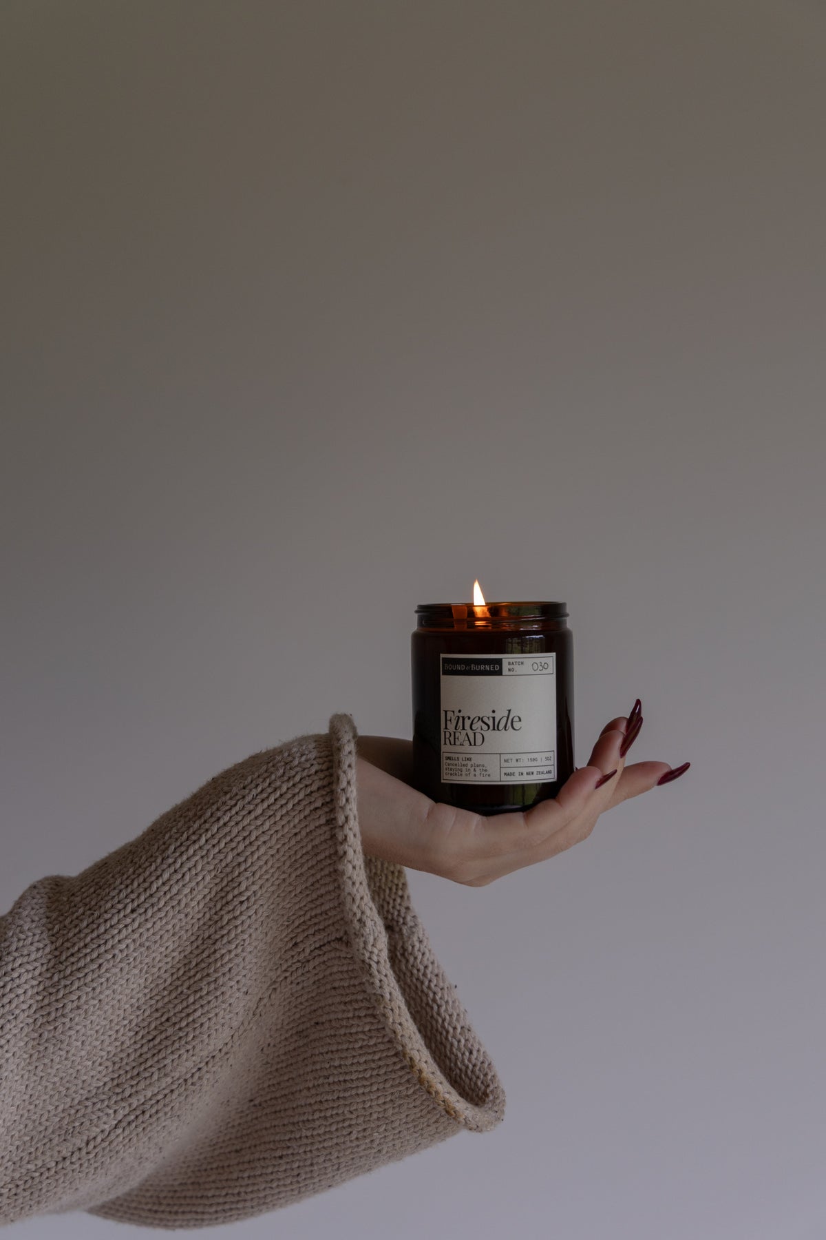 A lit candle in an amber glass jar held in a person's hand, wrapped in a beige knit sweater sleeve against a plain light gray background.