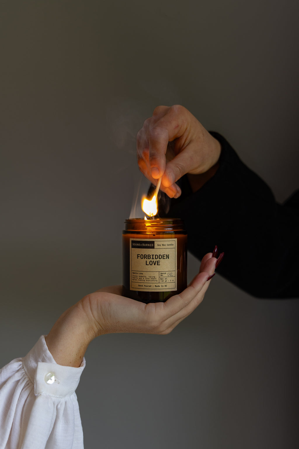 A person holds an amber glass candle jar while another hand uses a match to light the flame. The label reads 'FORBIDDEN LOVE'.