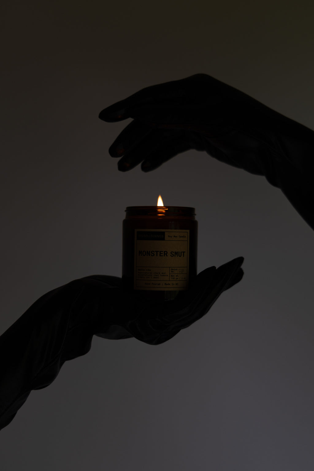A lit amber glass candle jar held between two dark silhouetted hands against a plain light gray background. The label text is mostly unreadable due to the low light.