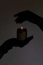 A lit amber glass candle jar held between two dark silhouetted hands against a plain light gray background. The label text is mostly unreadable due to the low light.