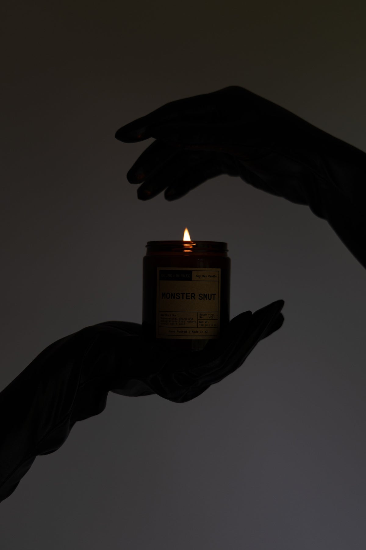 A lit amber glass candle jar held between two dark silhouetted hands against a plain light gray background. The label text is mostly unreadable due to the low light.