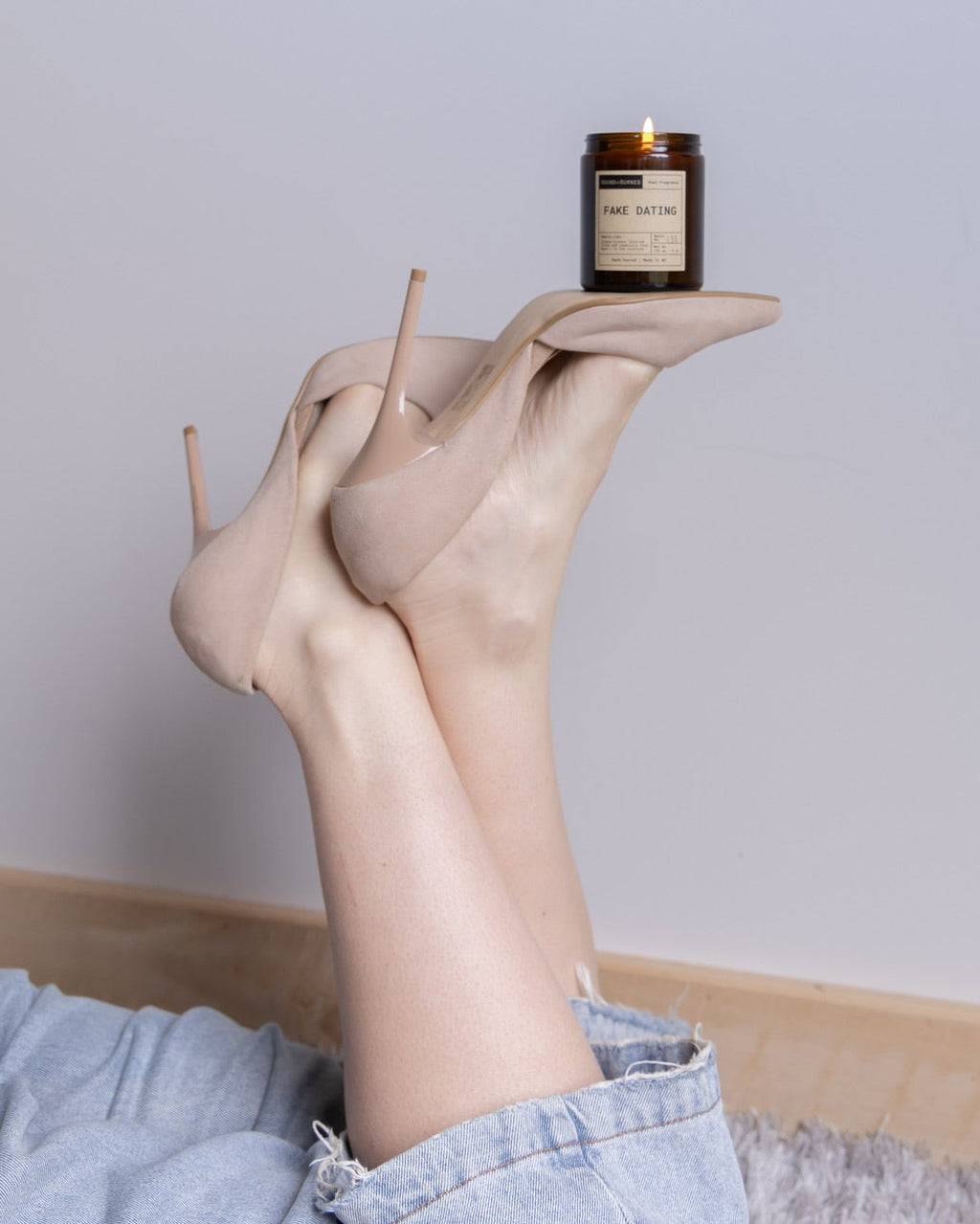 A person reclining on a light gray rug with their legs raised, wearing nude high-heeled shoes and light blue jeans. A small amber glass candle jar is balanced on the toes of the shoes against a plain light gray wall.