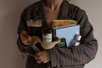 A person in a brown and black plaid blazer holds a curated bundle of items, including a glass of black coffee, a Coffee Shop Read candle, a notebook, a light blue book, and a perfume bottle, set against a plain beige wall.