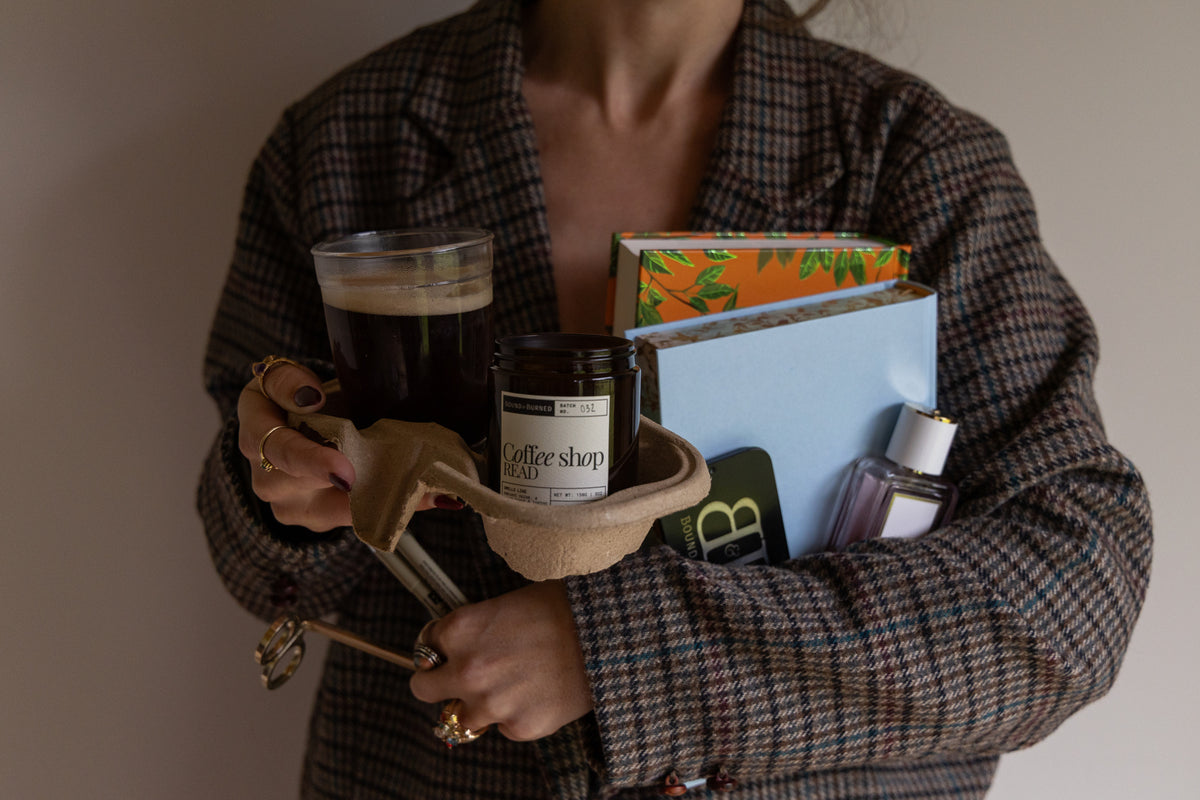A person in a brown and black plaid blazer holds a curated bundle of items, including a glass of black coffee, a Coffee Shop Read candle, a notebook, a light blue book, and a perfume bottle, set against a plain beige wall.