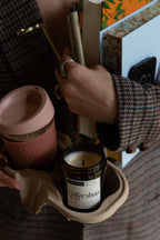 A person in a brown plaid sweater holds a reusable pink travel mug and a small amber glass candle labeled 'coffee shop read' in a cardboard holder, with books and stationery in the background.