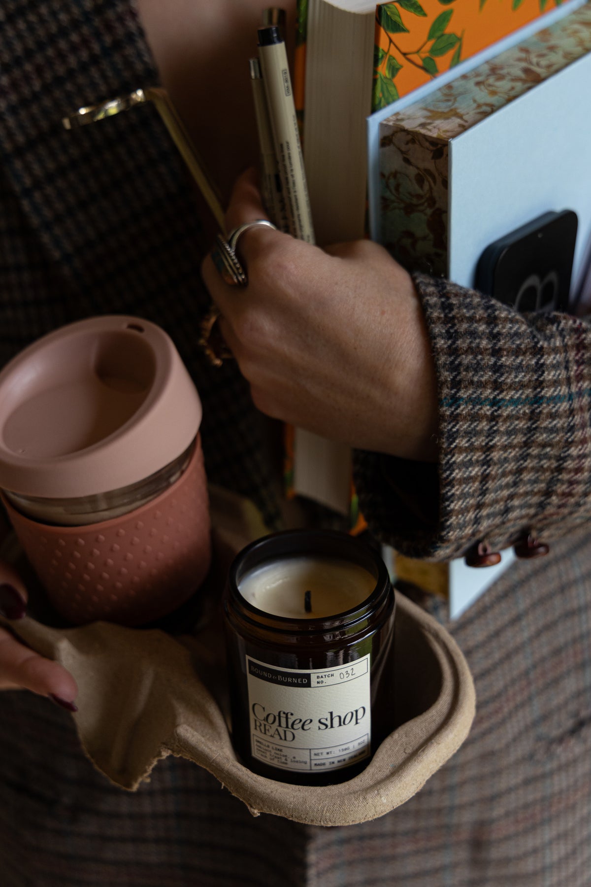 A person in a brown plaid sweater holds a reusable pink travel mug and a small amber glass candle labeled 'coffee shop read' in a cardboard holder, with books and stationery in the background.