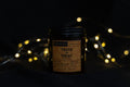 A dark glass jar candle with a black lid and a vintage-style beige label reading 'TRICK OR TREAT' and 'Spooky Scents', set against a dark background with warm glowing fairy lights.