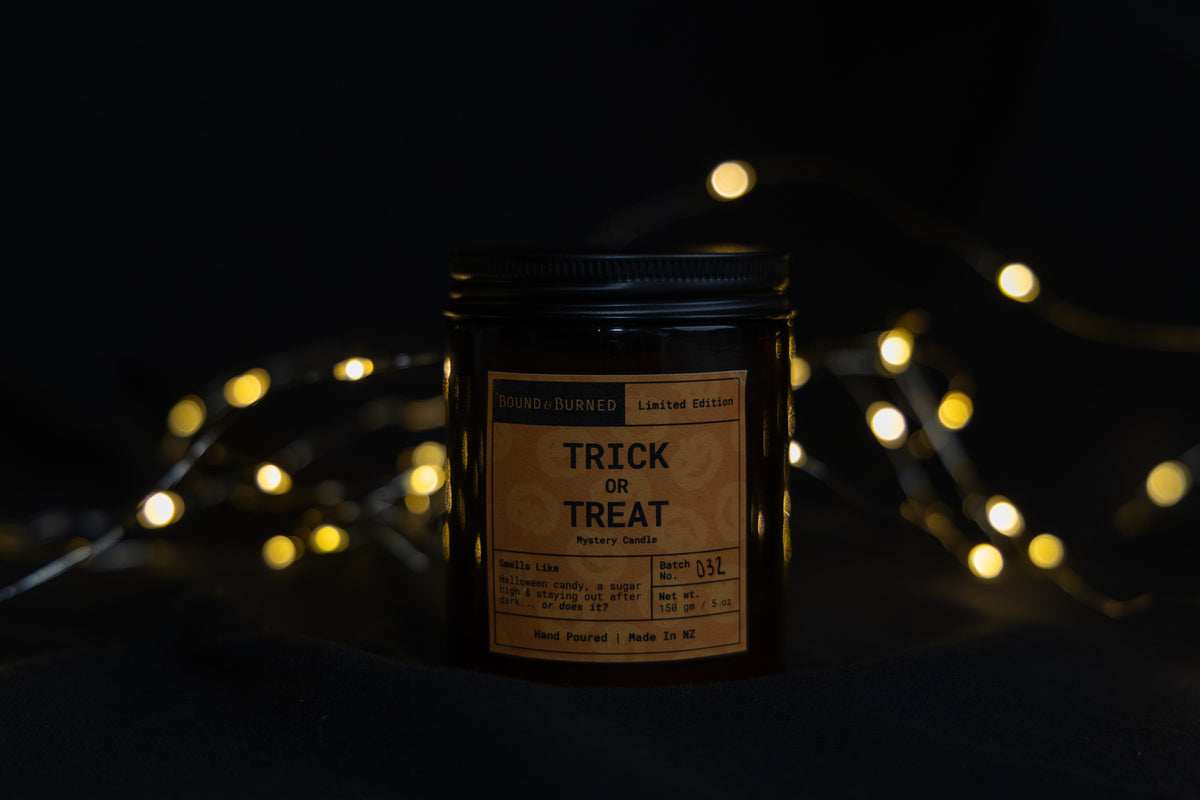 A dark glass jar candle with a black lid and a vintage-style beige label reading 'TRICK OR TREAT' and 'Spooky Scents', set against a dark background with warm glowing fairy lights.