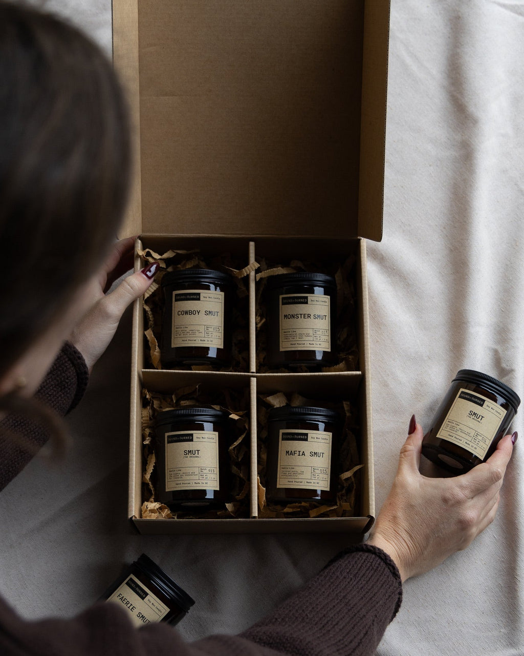 An overhead view of a person unpacking a cardboard gift box containing four dark glass jars with cream labels, placed on a white fabric surface.