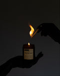 A dark, moody shot of a candle being lit. A hand holds a lit match above a small amber glass jar candle labeled 'MONSTER SMUT' against a dark, near-black background.