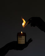 A dark, moody shot of a candle being lit. A hand holds a lit match above a small amber glass jar candle labeled 'MONSTER SMUT' against a dark, near-black background.