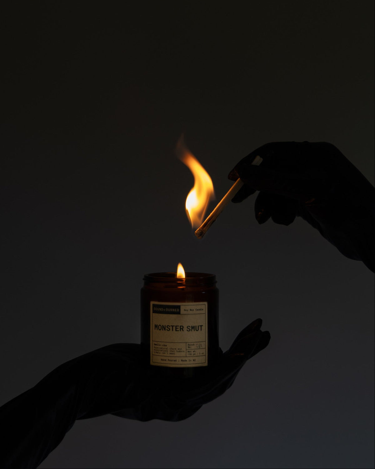 A dark, moody shot of a candle being lit. A hand holds a lit match above a small amber glass jar candle labeled 'MONSTER SMUT' against a dark, near-black background.