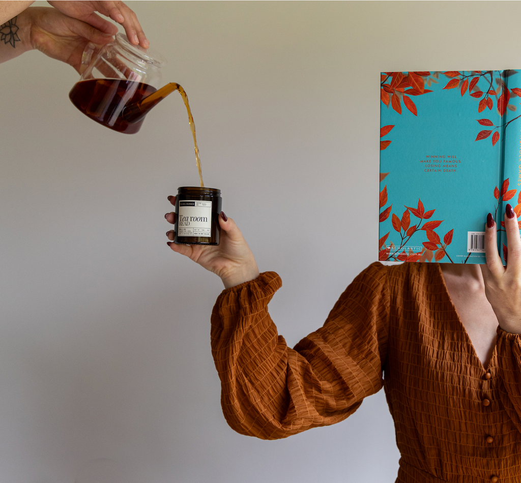 A person in a rust-colored crinkle blouse holds a small amber jar labeled 'Tea room read' while another hand pours tea from a glass teapot into it. A turquoise book with red-orange leaf illustrations covers the person's face.
